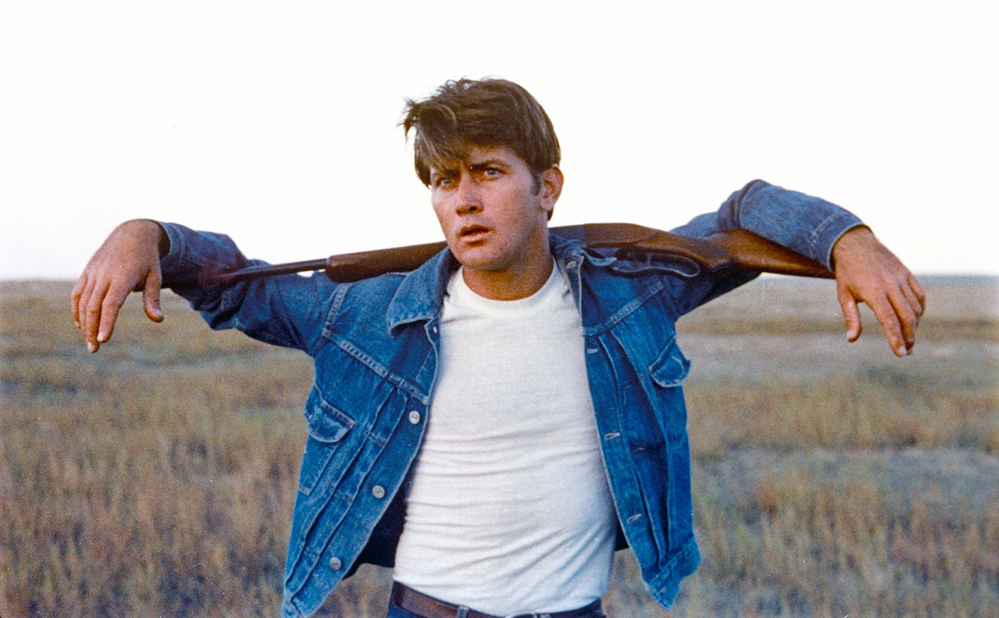 A young man in a jean jacket and white T-shirt stands in a field with a rifle across his shoulders