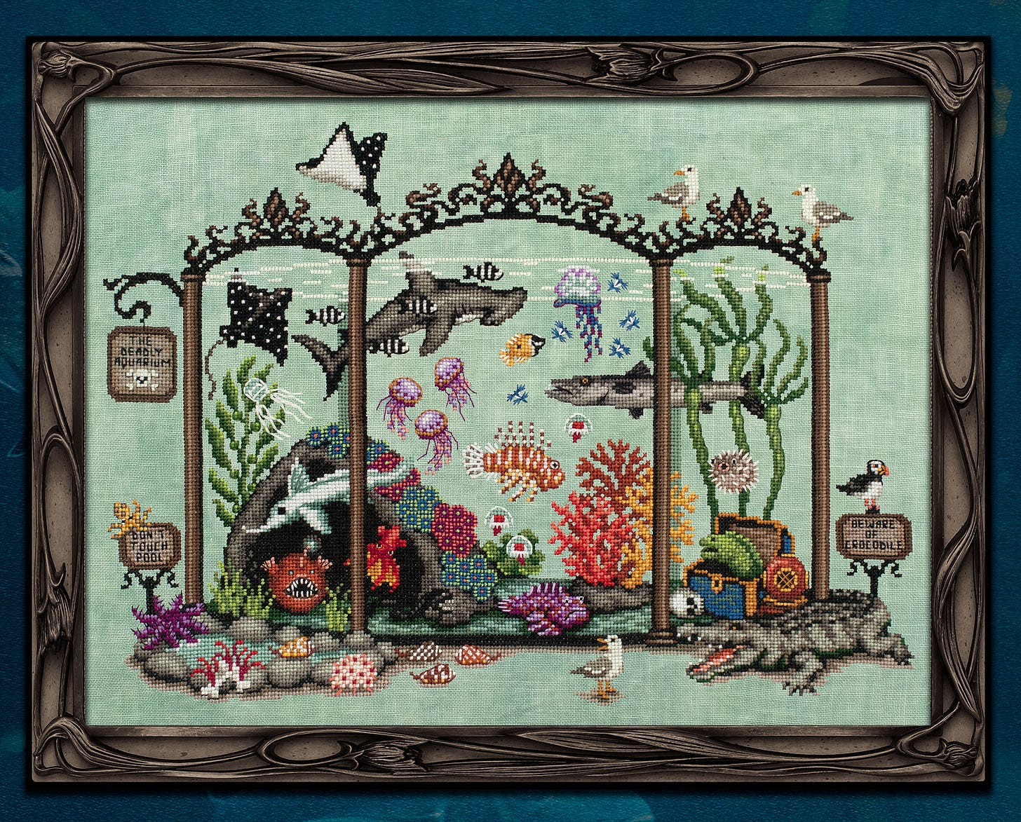 A framed cross stitch sampler depicting a glass aquarium hosting different corals, rays, and strange and dangerous sea creatures.