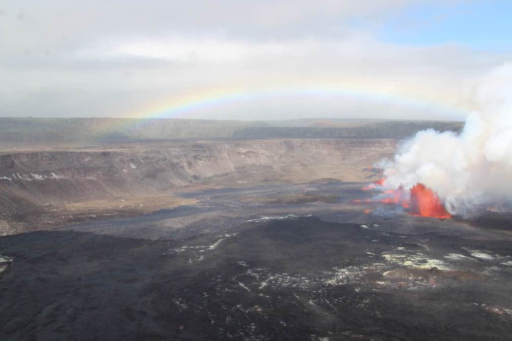 Bright rainbow over a large volcanic crater as a plume of lava erupts from one side producing a cloud of thick white smoke.