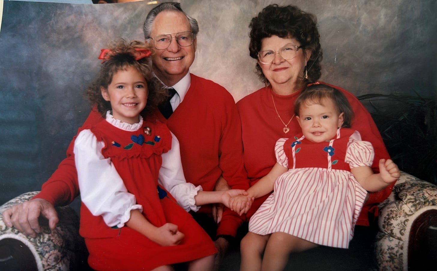 A 6 year old and two year old girl sit on their grandparents' laps, holding hands. Everyone is wearing red and smiling.