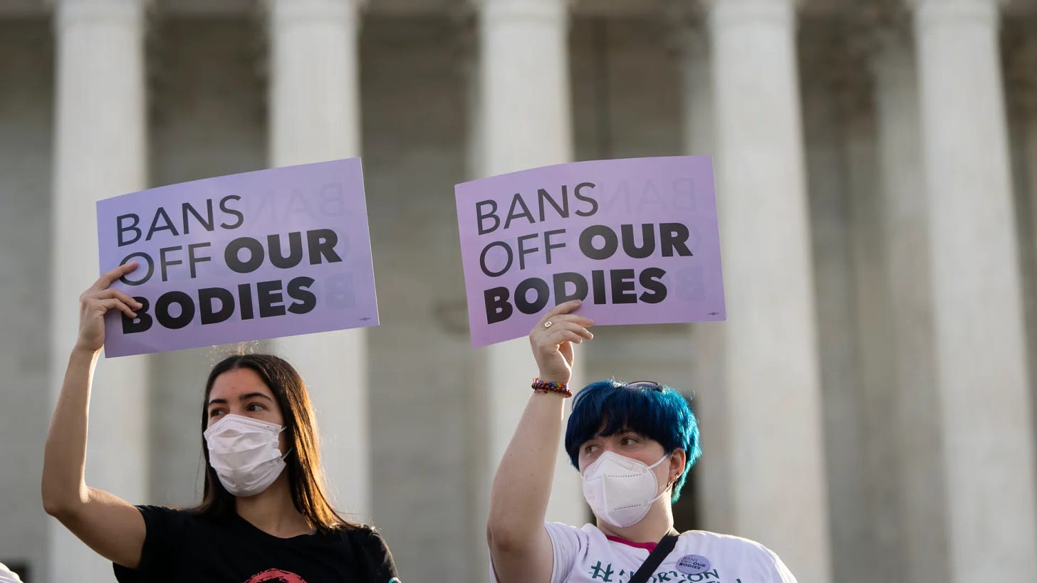 A photo of two protestors wearing masks holding up purple signs that read, “BANS OFF OUR BODIES.”