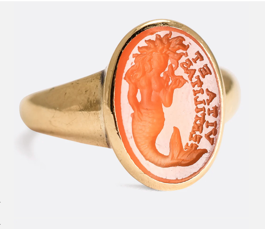 gold ring with large orange oval stone, engraving depicts a mermaid, with long hair and detailed tail and next to it are the words "Vita et Fertilitas", translating to "Life and Fertility"