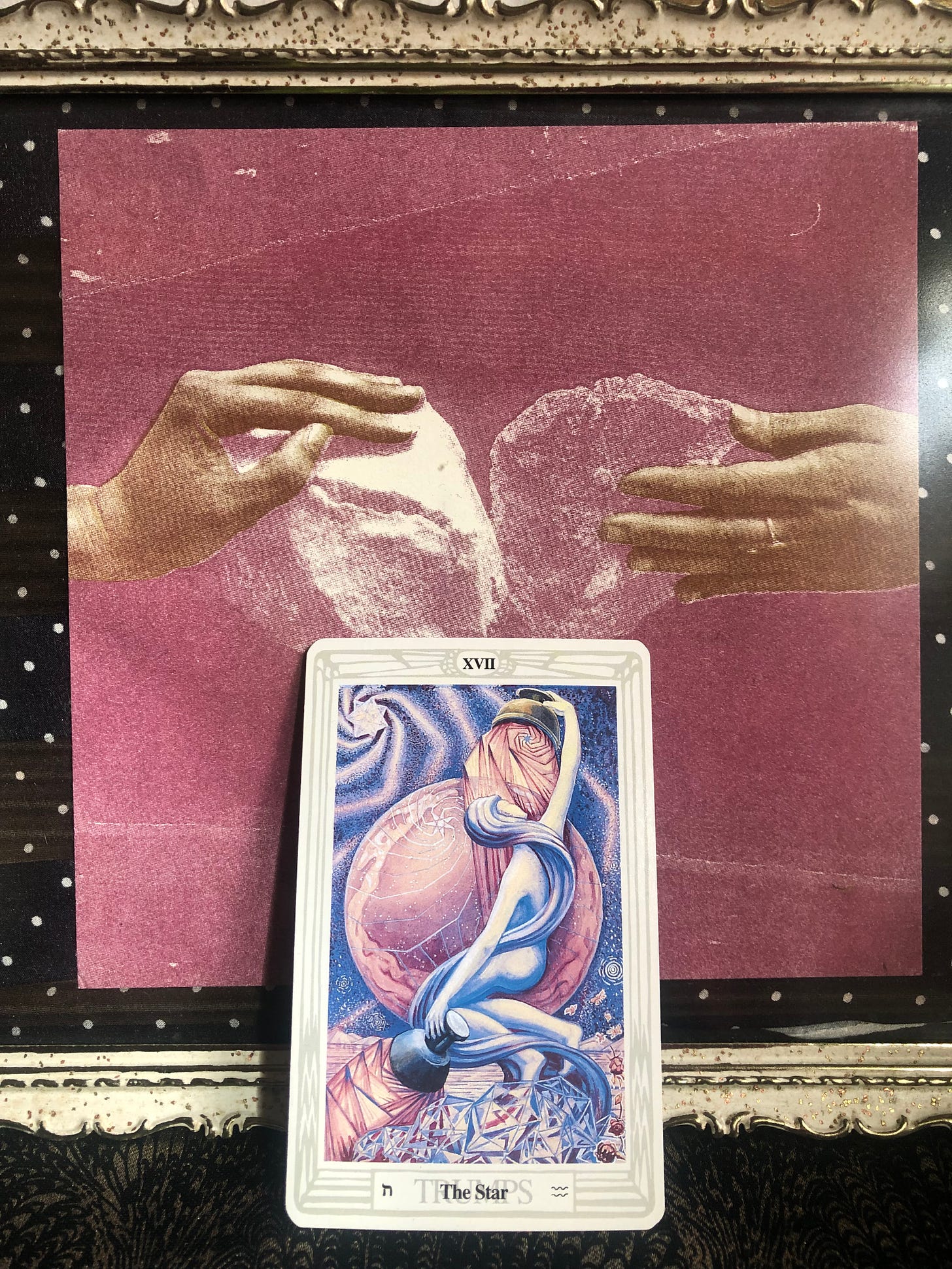the star tarot card from the thoth deck rests against a framed art piece of two hands each holding a stone with the two stones touching in the center. 
