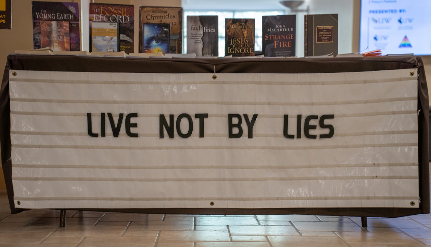 A white table banner reads "LIVE NOT BY LIES" in removable black letters. Young earth creationist literature adorns the top of the table.