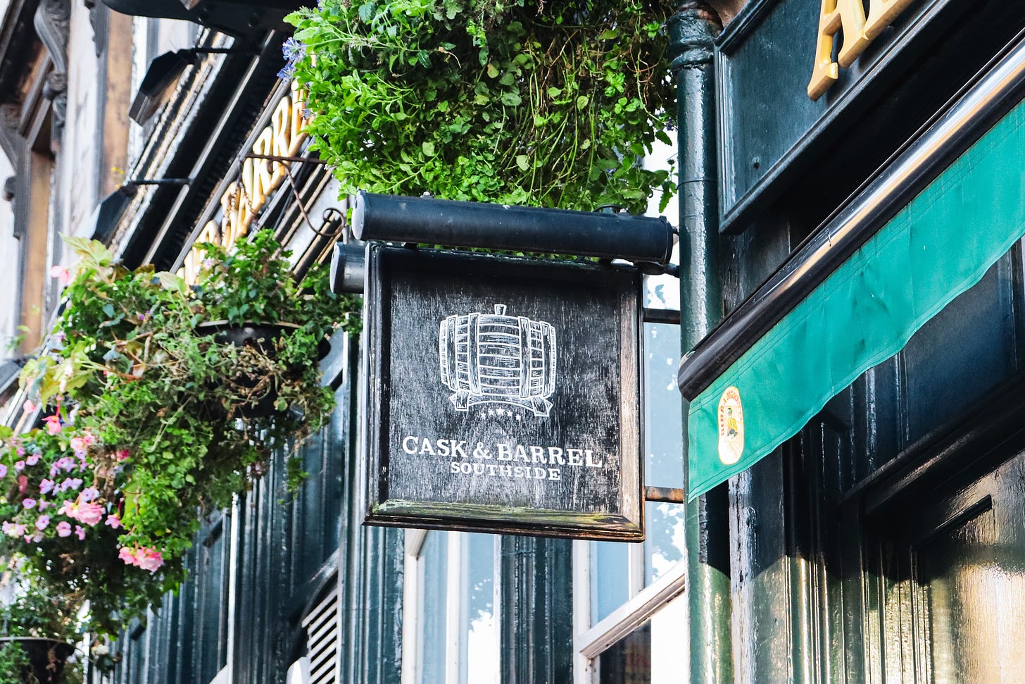 The sign of Cask and Barrel Southside, white chalk effect on black board. With hanging baskets behind.