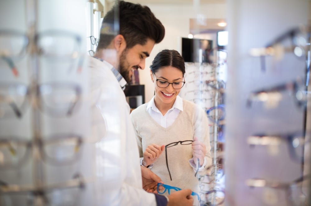 A smiling woman in a beige sweater holds a pair of glasses while talking with an optician in a white coat. They are standing in an optical store, surrounded by displays of eyeglass frames. The optician is showing her different frame options.