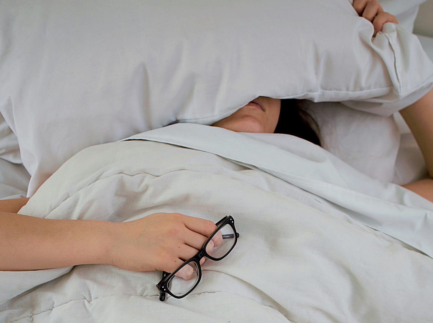 woman lying in bed covering face with pillow and holding pair of glasses