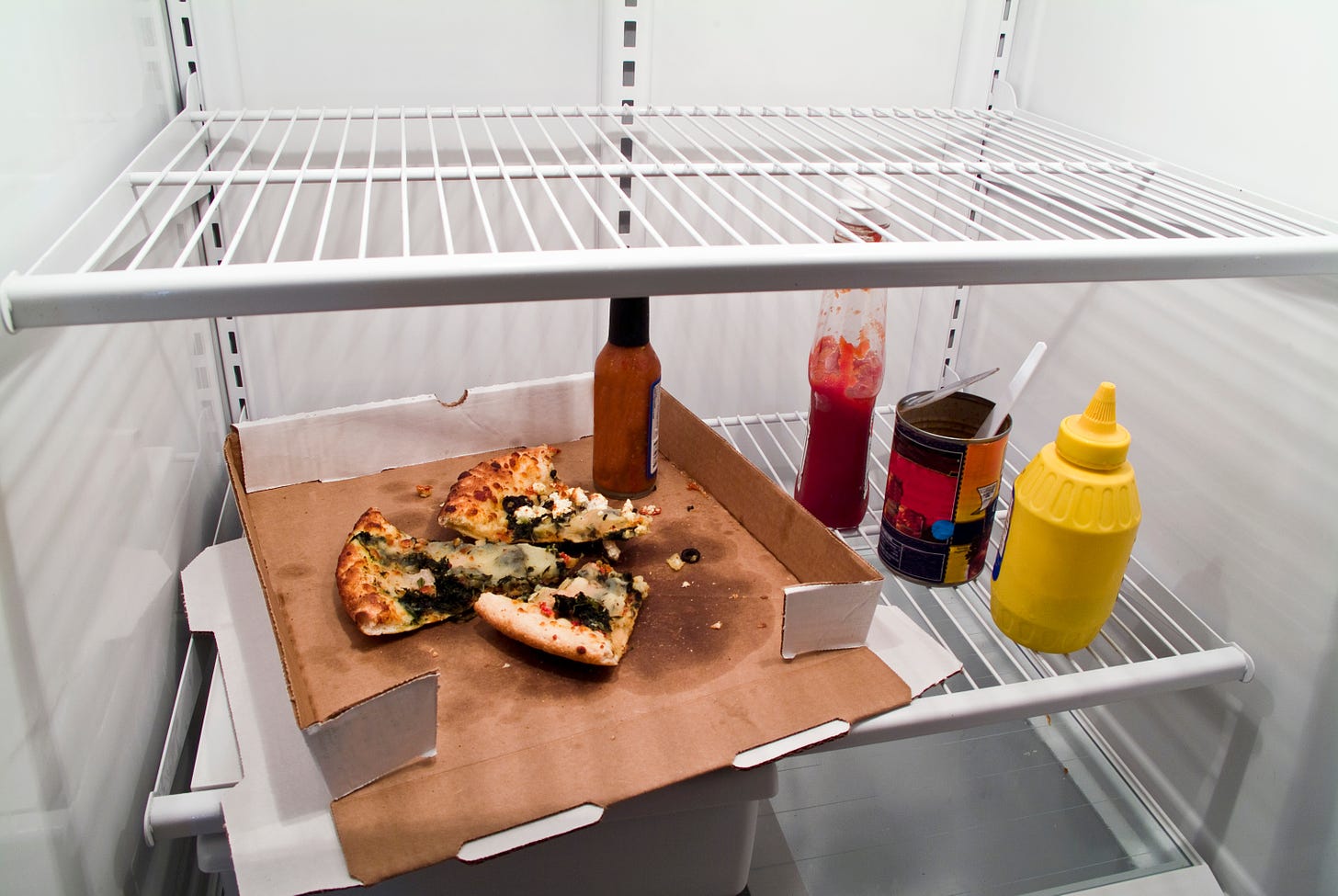 Mostly empty refrigerator with a pizza box inside containing a few slices of pizza; next to it our some condiments