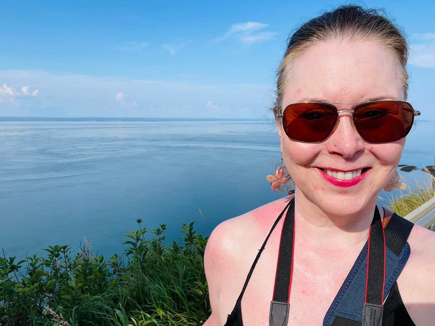 A white woman with a slight sunburn and wearing sunglasses, a black halter top, a camera around her neck, and hair in a ponytail stands on a platform on a cliff over looking the ocean.