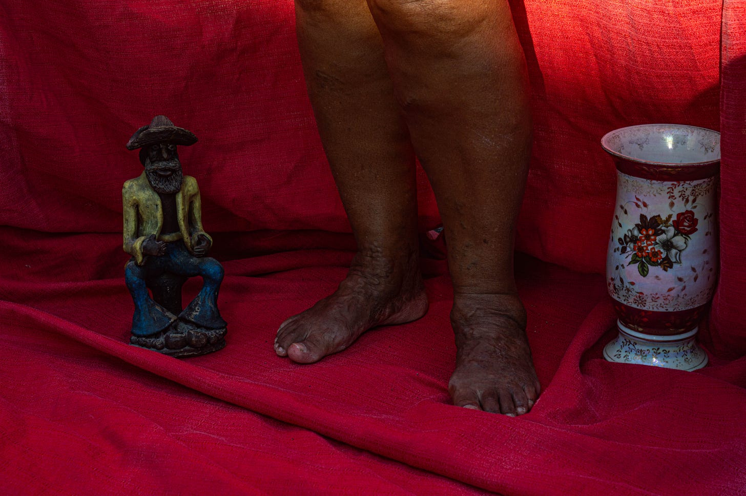 a small statue of a man sitting next to a pair of feet. On the right side of the feet is a black and red vase. The ground is covered with red cloth