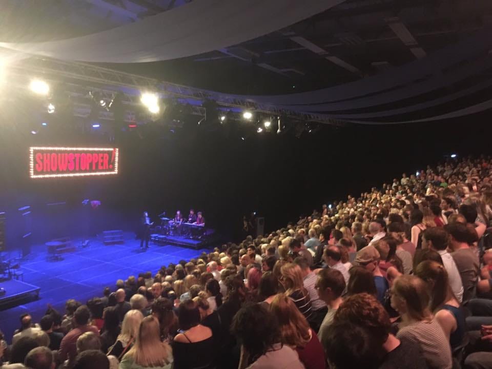 photo taken from the back on a big room over the heads of loads of audience members looking down onto a wide stage where a man is talking and a red sign with "Showstopper" hangs on the back wall. To the left of the man are three musicians on keyobard, percussion and wind instrument