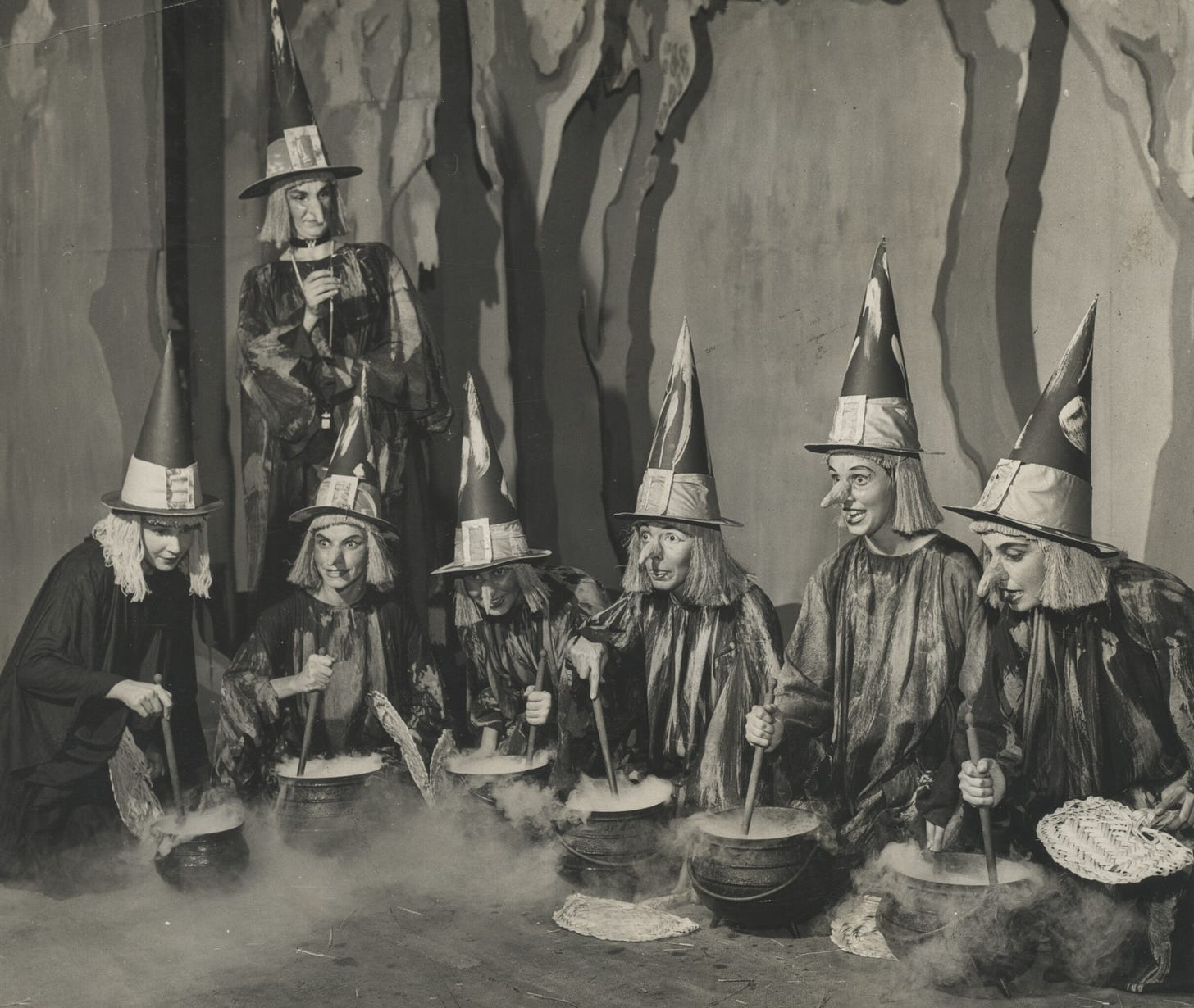 Black-and-white photo of young women in witch costumes (complete with long fake noses) stirring steaming cauldrons.