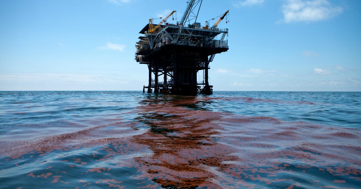 An oil platform floats under a blue sky in the background. In the foreground, reddish-brown crude oil floats on the surface of the water.