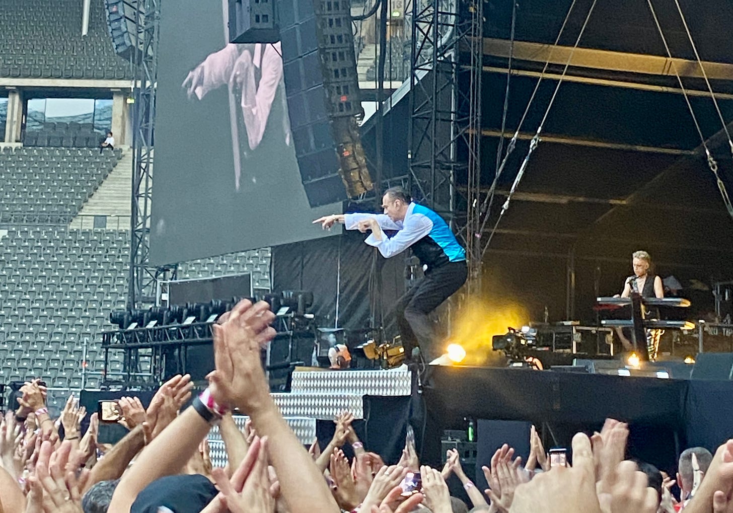 Imagem: Dave Gaham cantando na ponta do palco em frente a uma escada. Braços do público levantado e uma distância de 10 metros do palco.