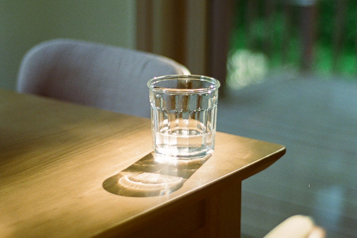 Glass of water sits on a wooden table, half-full. Light from a window casts a shadow of the glass on the face of the table.
