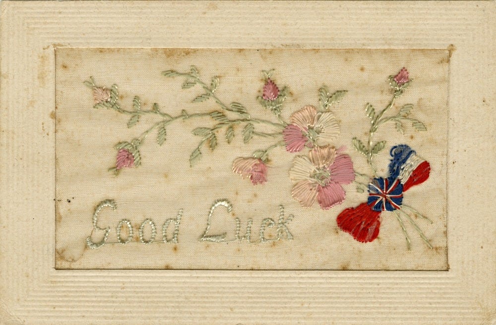 A pale pink sil postcard embroidered with delicate flowers, a mini flag, and the words 'Good Luck'