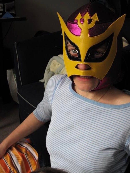 Your humble correspondent in a stripped blue and white shirt, and wearing a yellow leather-look and shiny pink lucha libre (Mexican masked wrestling). As well as black rimmed eyes and nose, the mask features pointy cat ears, as it is the mask of lucha libre character El Felino.  This photo does not represent the author's actual costume, which involves an N95 mask.