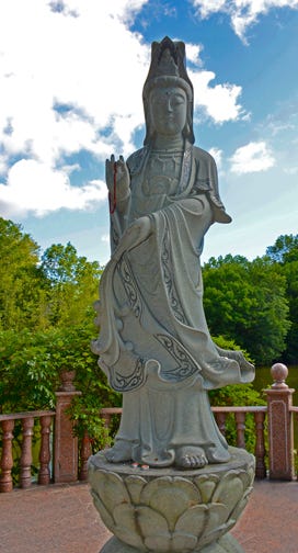 A statue of the god/dess Quan Yin in Kent England stands atop of lotus blossom. S/he holds one hand in front, palm up, and one hand in front as if to stop something.