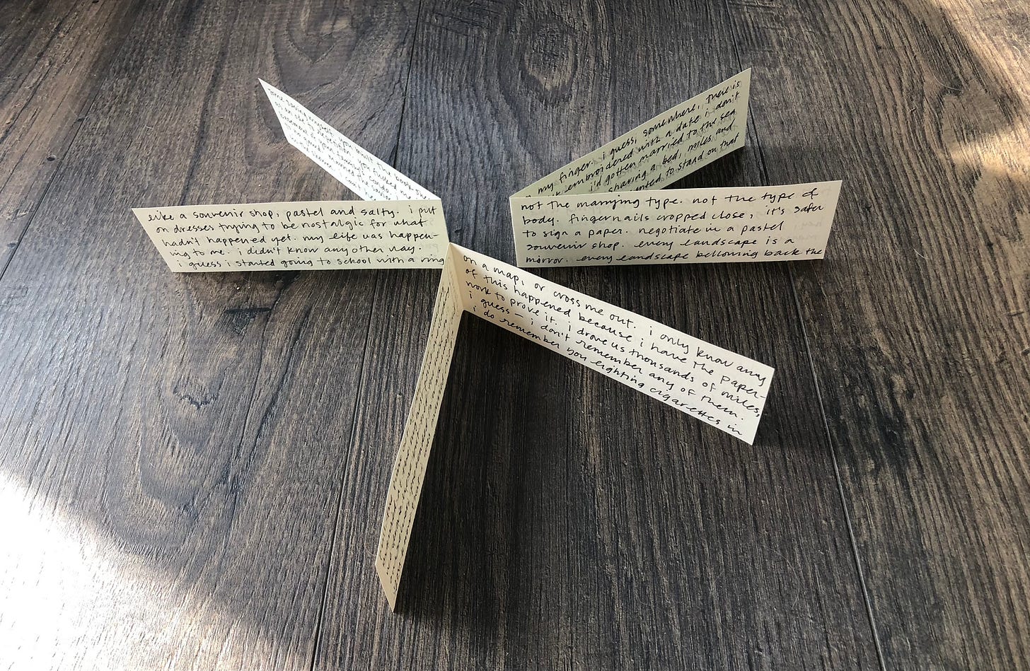three folded scraps of paper with black ink handwriting standing in a circle on a wooden floor