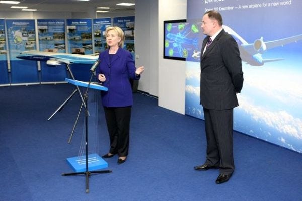 A blonde-haired woman in a blue coat and black pants, behind a podium, gesturing towards a taller man in a dark gray suit standing in front of a picture of a jet airliner