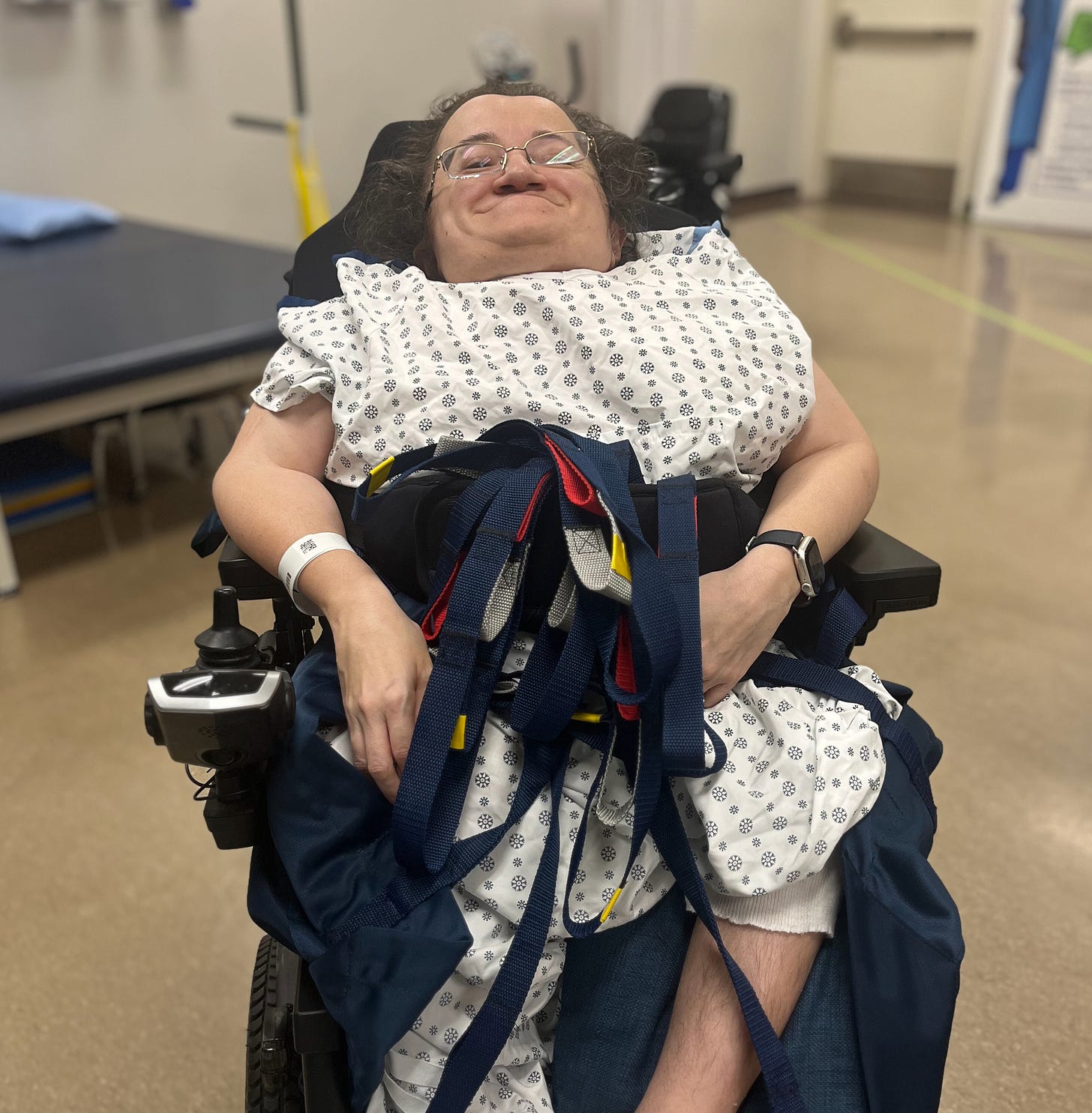 Photo of Athena sitting in her power wheelchair. She's smiling in her white hospital gown with the straps of the hoist sling around her waist.