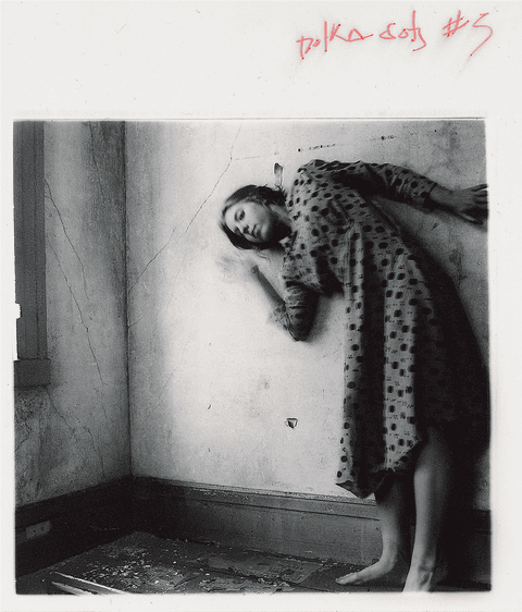 Polka Dots #5 by Francesca Woodman, 1976. In a grainy black and white image, a woman in a long sleeved dark polka dotted dress leans at almost an angle, one arm thrown away from her, the other curved up as though she about to stretch, clasp her cheek or fling her arm upwards quickly. It’s as if quick movement has been captured and pinned down on the page.