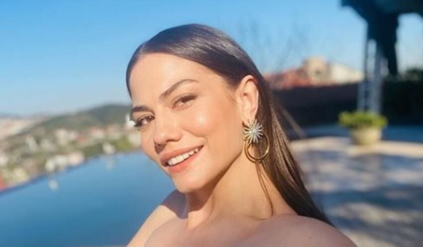 İstanbul Boğazı manzaralı bir terastan güneşli bir günde gülümseyen Demet Özdemir. Uzun düz saçları, bronz teni ve altın renkli küpeleriyle kameraya bakıyor.
