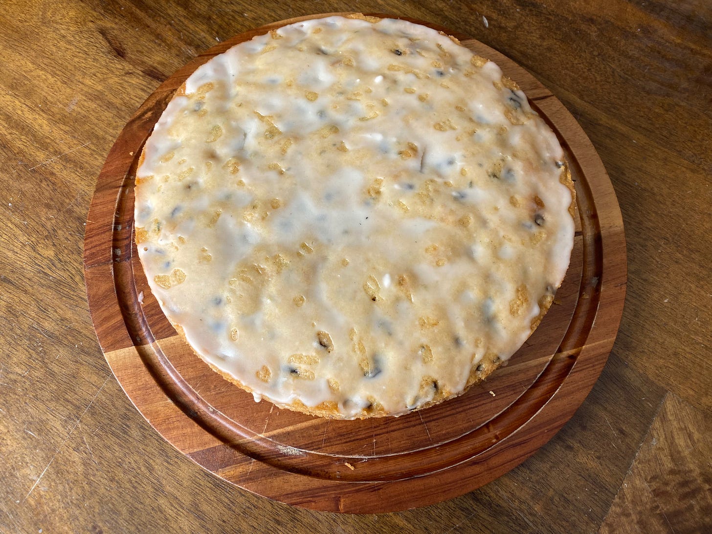 A round cake drizzled  with a thin lemon glaze and studded with specks of rosemary sits on a wooden cake board.
