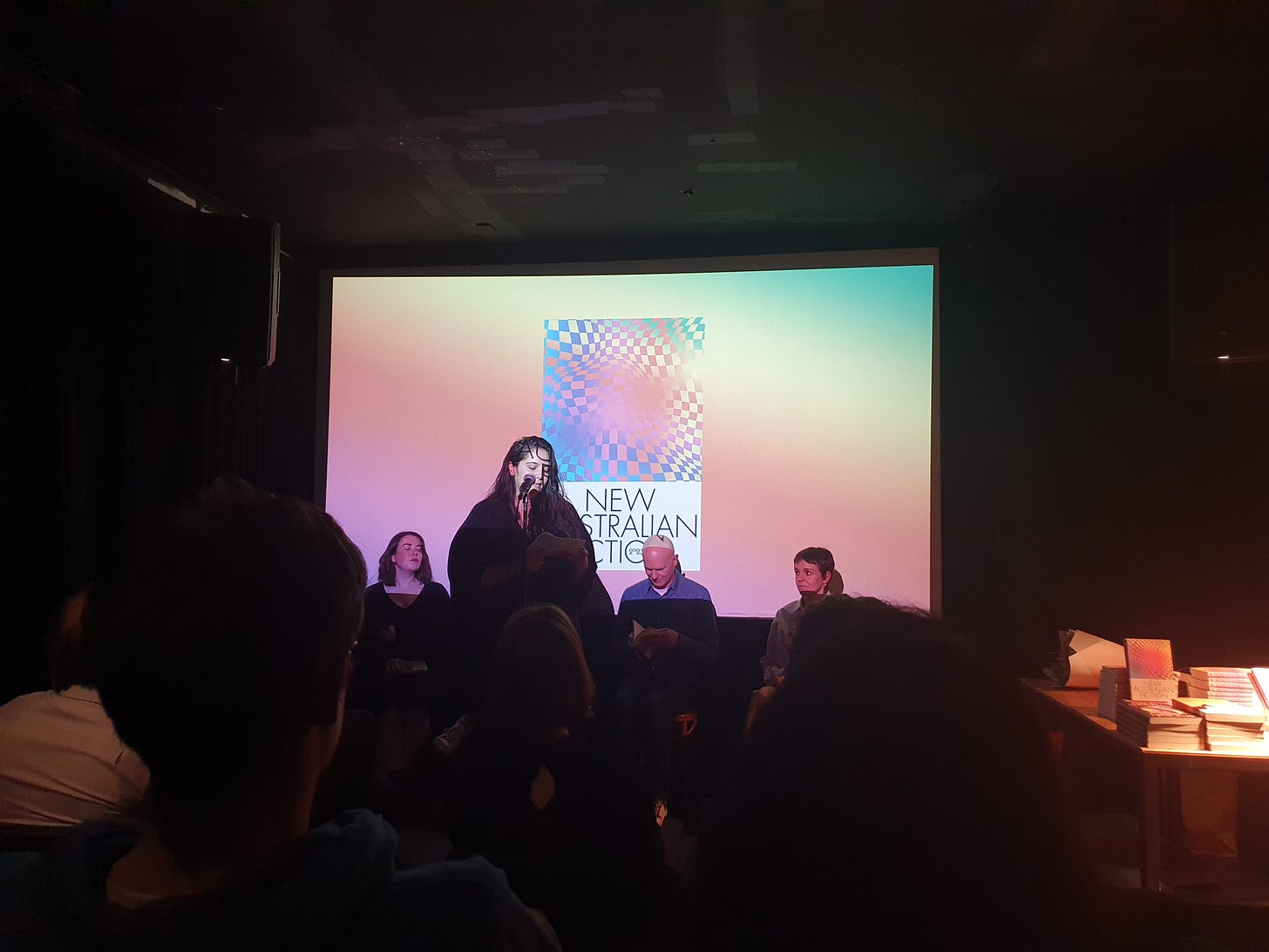 A woman standing a a microphone, speaking to an audience. Behind her is a panel of seated people and a projected image of the New Australian Fiction 2023 cover