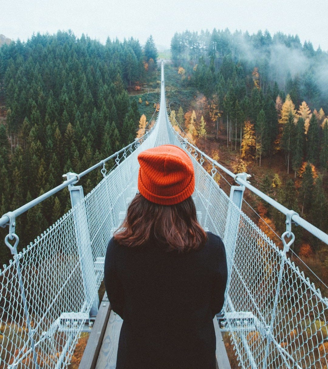 woman wearing knit cap walking on white bridge between trees during daytime woman wearing knit cap walking on white bridge between trees during daytime