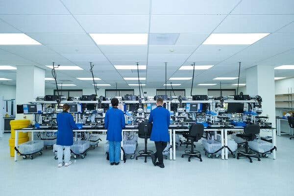 Workers in blue lab coats are seen from the back as they stand at a row of tables packed with lab equipment.