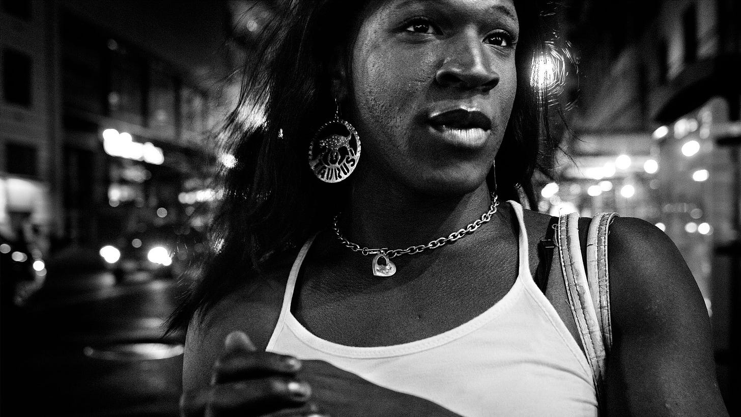 A high-contrast black-and-white photograph of a Black woman wearing a white tank top, a thick chain necklace and big Taurus earrings. She looks off camera while standing on a city street at night.