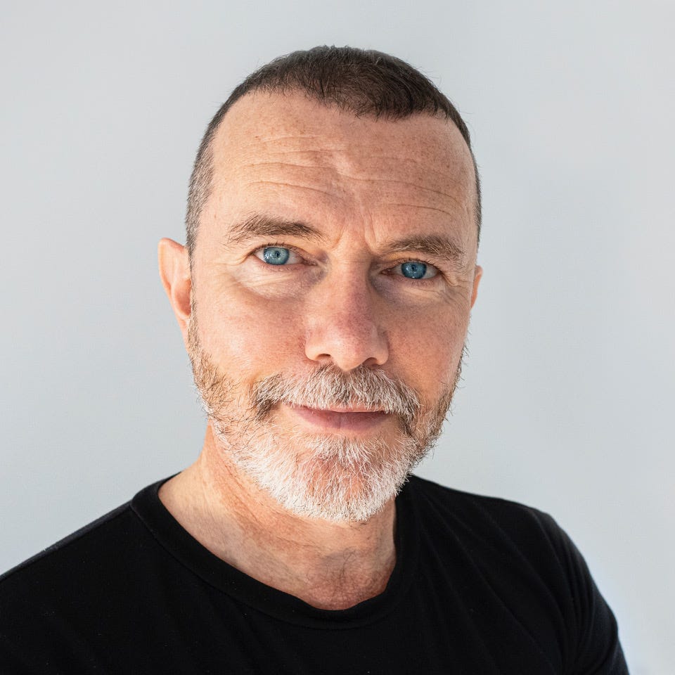 Joe is a white man with very short brown hair, blue eyes, and a grey beard. In this photo, he’s standing in front of a grey backdrop wearing a plain black t-shirt. He is staring straight into the camera, smiling ever so slightly with his mouth closed. 