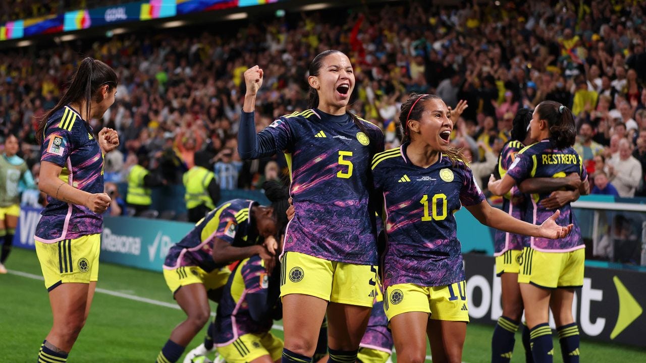 In a crowded soccer stadium, a group of women from the Colombian soccer team are celebrating a goal, with two of them in the foreground smiling shouting as they pump their outer arms and have their other arms around each other.