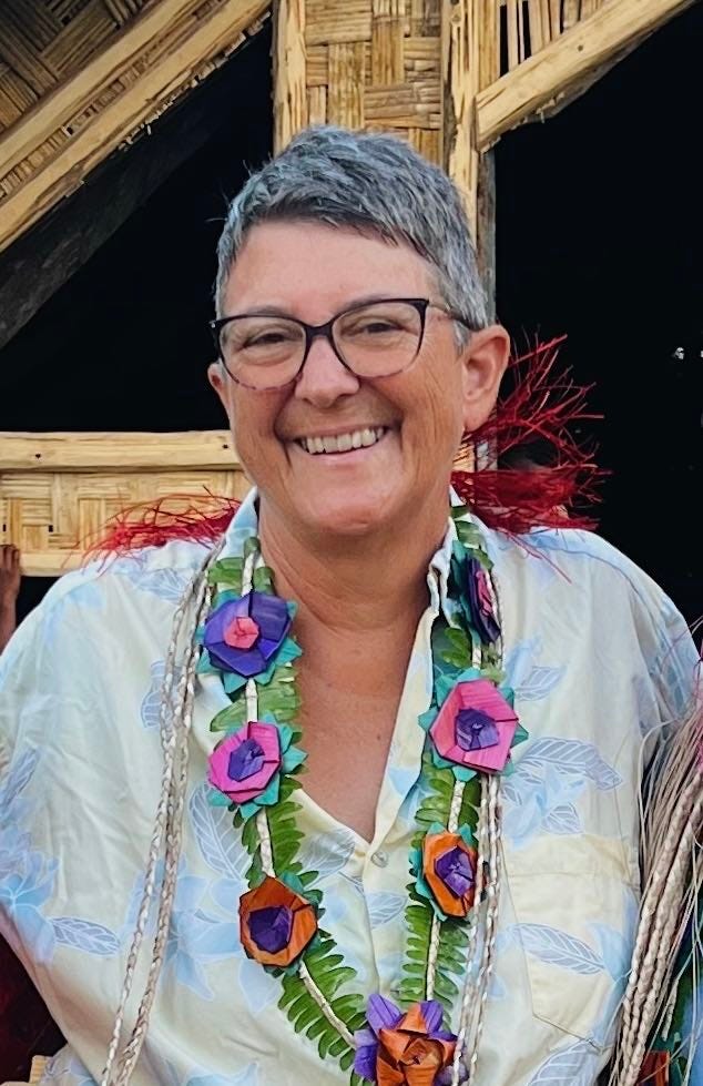 a middle aged woman standing in front of a thatch hut, wearing a lei of flowers