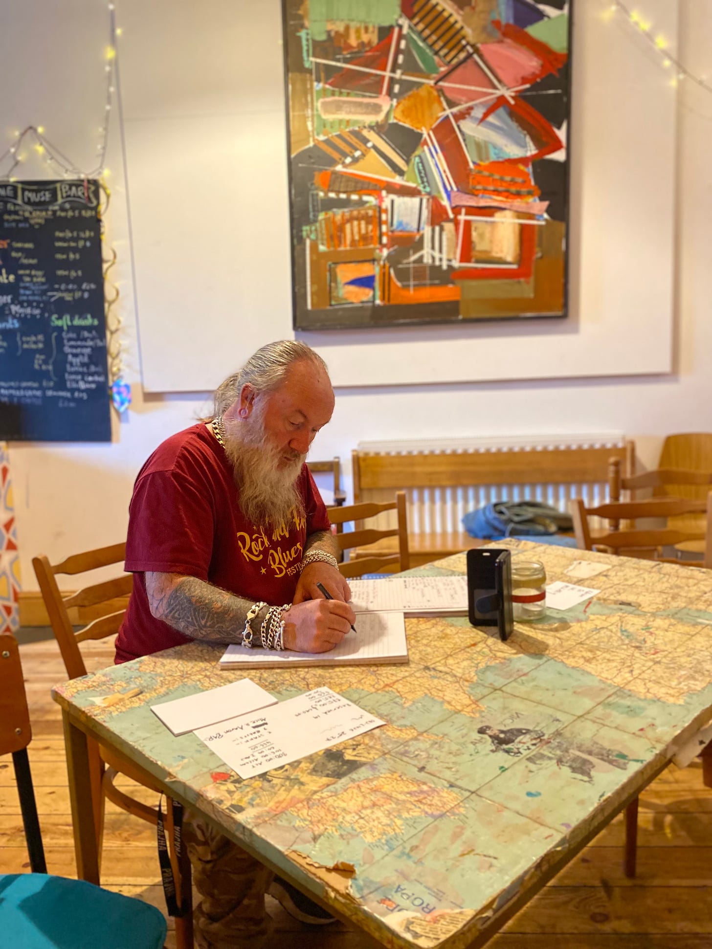 Man with beard sat at table writing. He has tattoos on his arm and wearing silver chains on his wrists. Painting on the wall in the background