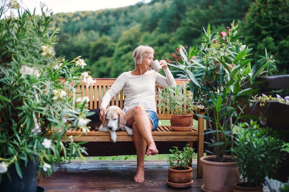 Elder woman sitting on a patio bench with her dog while reflecting on her day surrounded by plants.