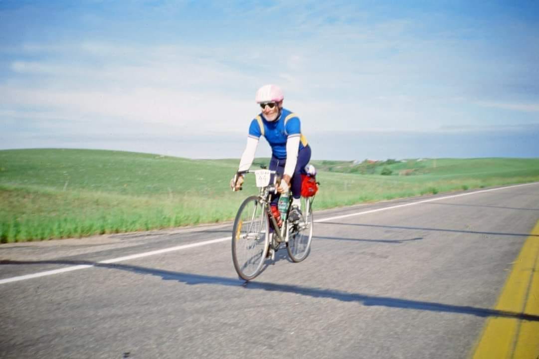 man with white beard, pink helmet, blue shirt, white sleeves, and navy pants rides a touring bicycle with skinny wheels and a red pack on the back atop a cement road with yellow dividing lines to the right and to the left and behind green grass and a blue and white striped sky