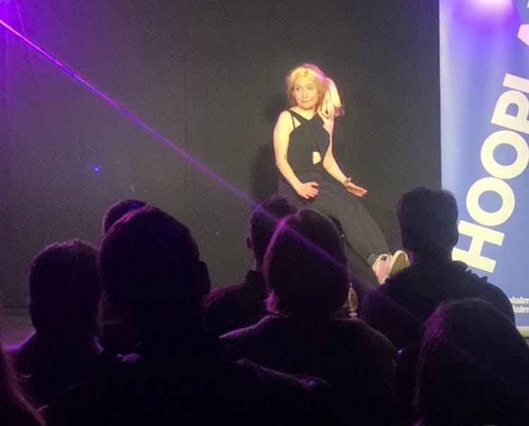 Woman sits awkwardly on high stool on a stage. She wears a blonde wig and looks out at the audience