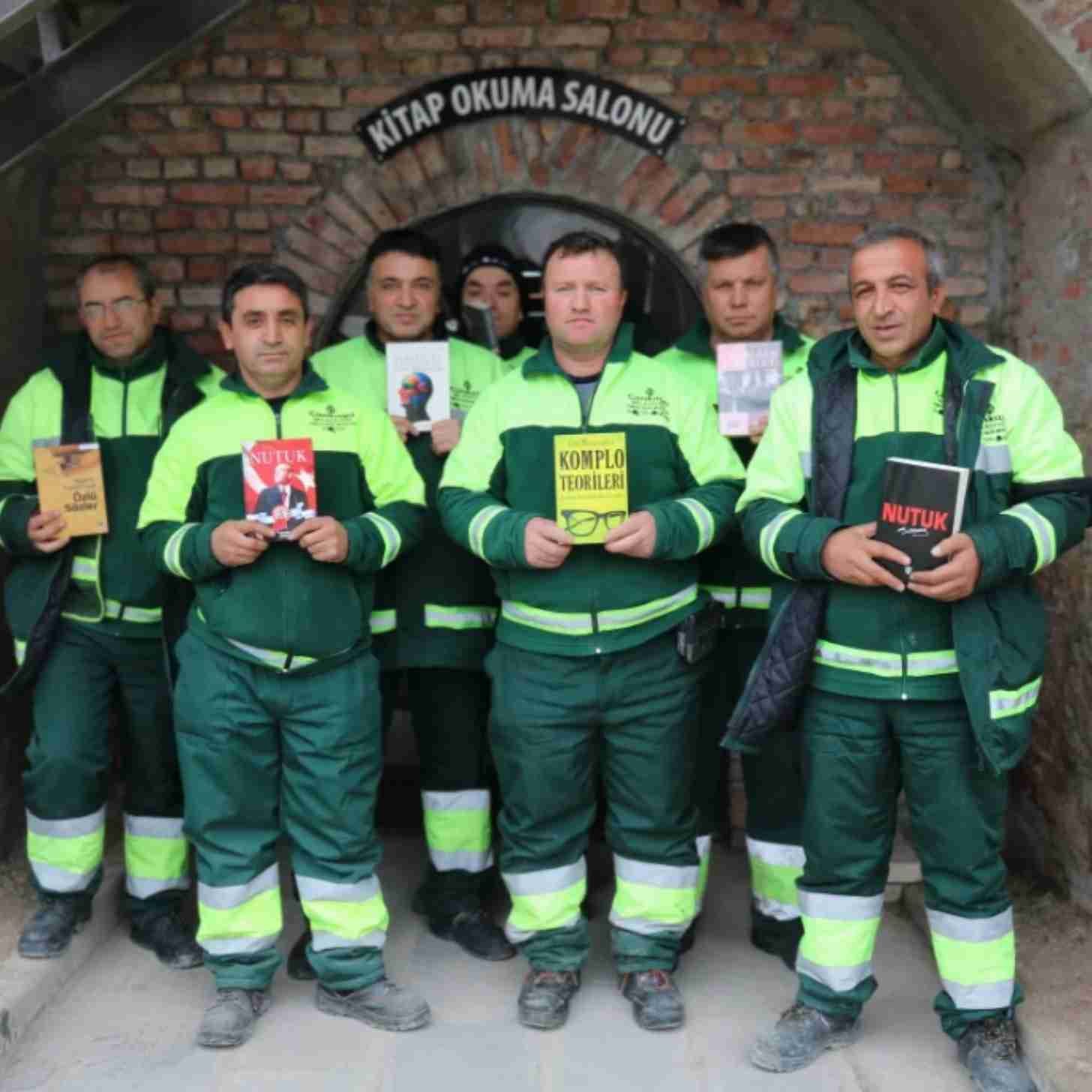 A group of male sanitation workers stand in front of a brick-facade entrance to a public library. They're all holding up books in their hands. 