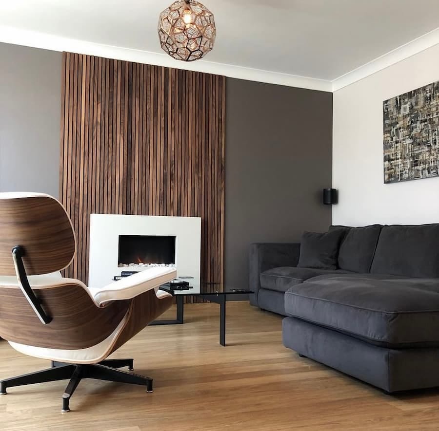 A modern living room with a fireplace and a wooden accent wall. The room has a wooden floor and a grey corner sofa with a throw and pillows. There is a white armchair with a wooden base and a coffee table with black legs and a glass top in front of the sofa. The fireplace is white and built into the wooden accent wall. There is a brown and white abstract painting hanging above the sofa. There is a decorative copper light pendant hanging from the ceiling.