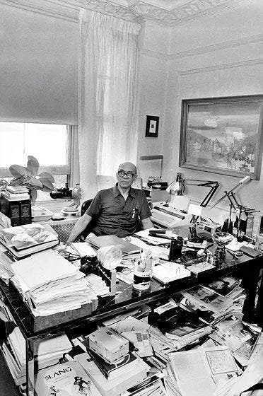 This may contain: black and white photograph of man sitting at desk covered with papers, books, and other items