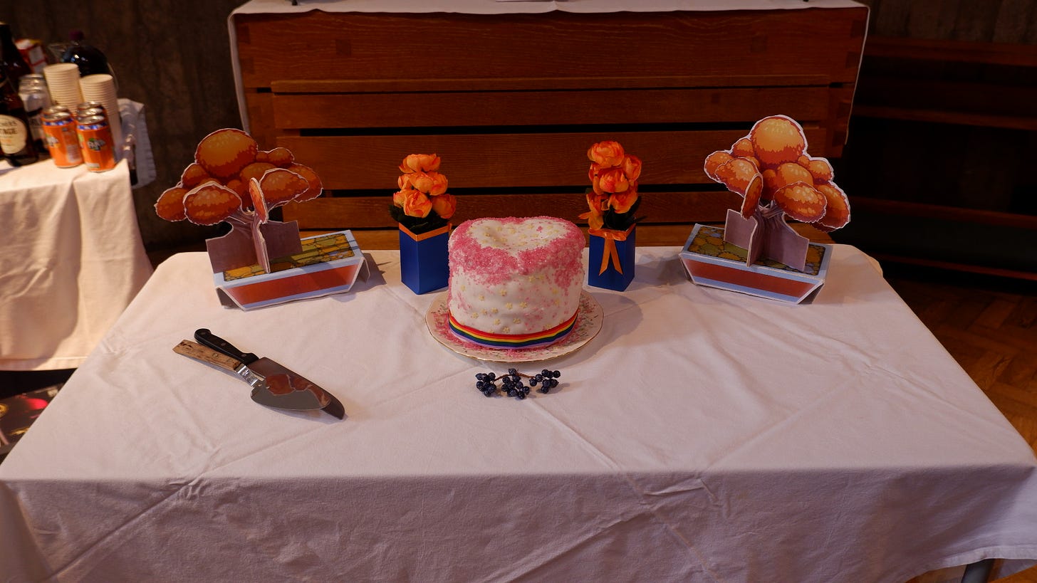 A table with a white tablecloth. On it sits a cake knife, a tree on either side, a blue and orange flower centrepiece on either side, and in the centre is a heart-shaped cake, white with a pride-coloured ribbon around the bottom.