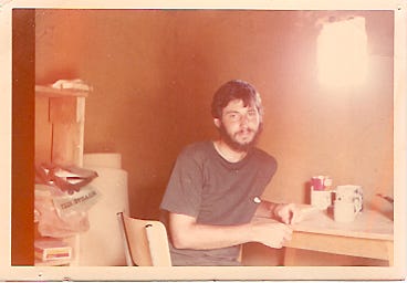 Photo of a 21 year old me sitting at a small table inside a small, adobe, thatch-roofed hut. 