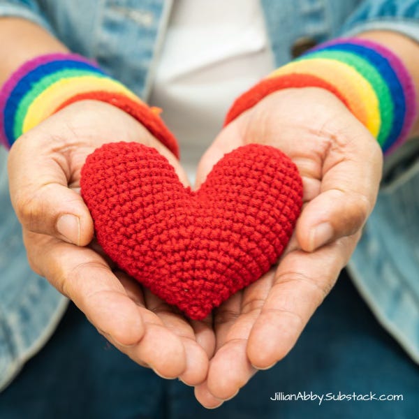 Hands with rainbow wrist bands holding a red, crocheted heart.