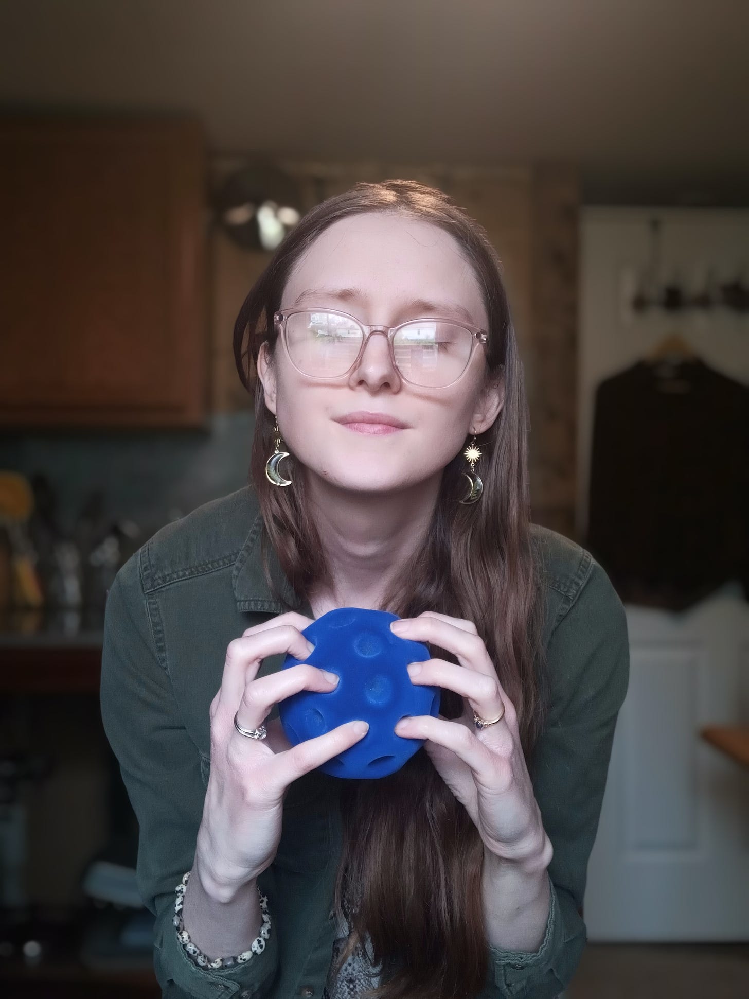 Kandi Zeller (she/her), a white woman with long reddish brown hair and pink glasses, holds a blue Rubbabu stress ball that looks like an oversized golf ball. Kandi has her eyes shut and appears calm.