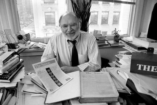 In a black and white photo, a balding, bespectacled Mr. Navasky, in a loosened black tie and rolled-up sleeves, at his desk, which is strewn with books, papers and a copy of The Nation.