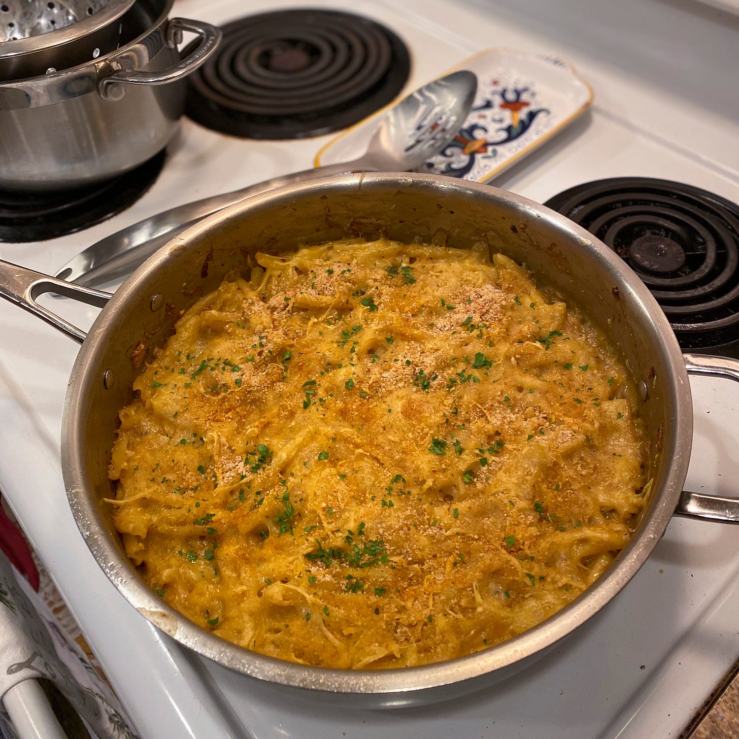 A large steel pan full of the mac & cheese described above, with browned spots of cheese and breadcrumbs on the top, and dusted with parsley. It sits on the stove next to an empty pot and a slotted metal spoon.