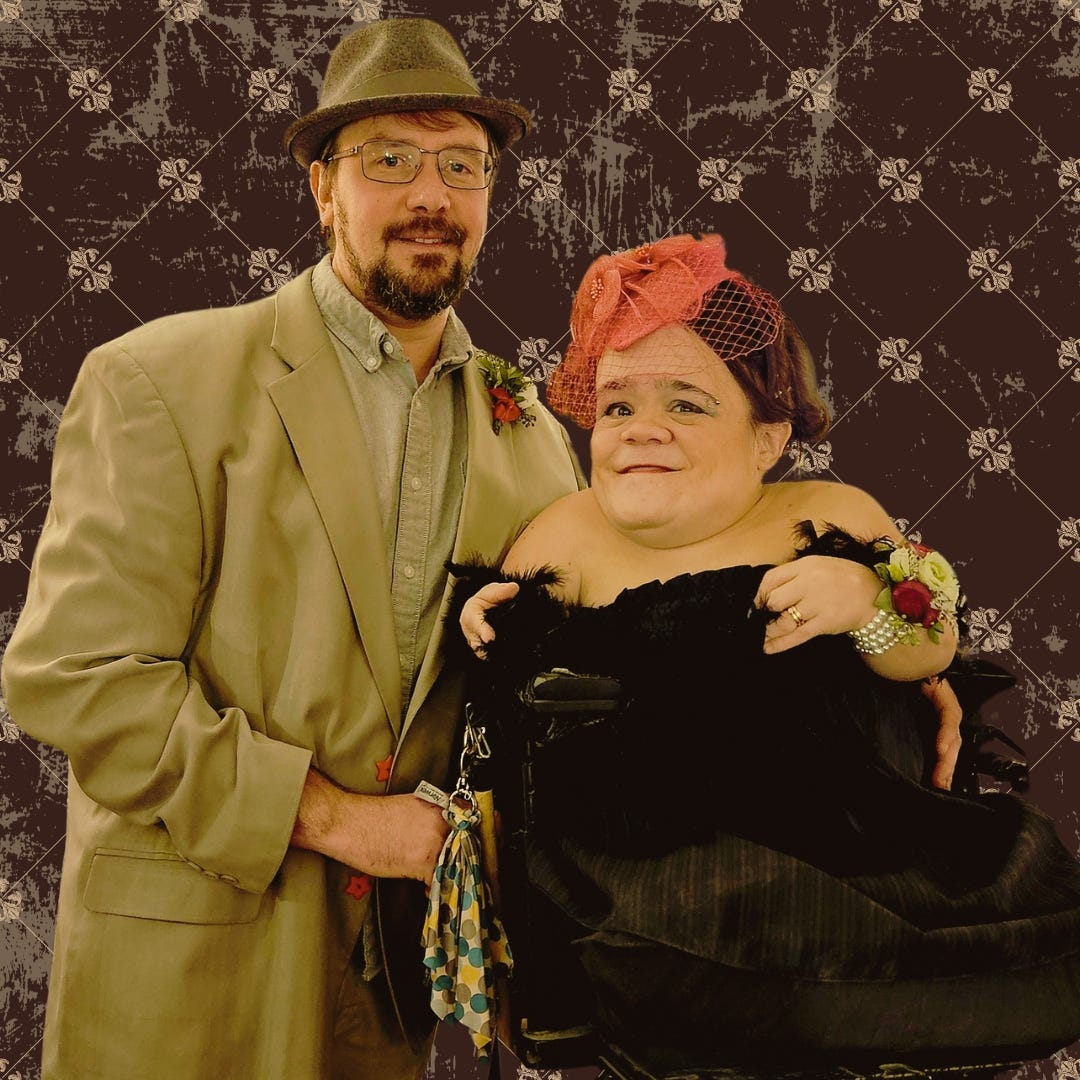 Gaelynn is wearing a black hat and a pink fascinator hat, and Paul is wearing a tan suit and a grey, wool hat with a brim.