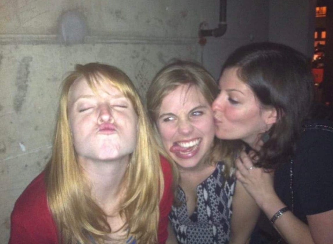 Three women making goofy faces next to the cement wall of a bar somewhere